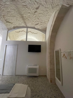 Ein Zimmer mit Steindecke, einem flachbildfernseher an der Wand, einer kleinen Klimaanlage, einem Bett mit Decke und einem Fenster mit Blick auf den Himmel.