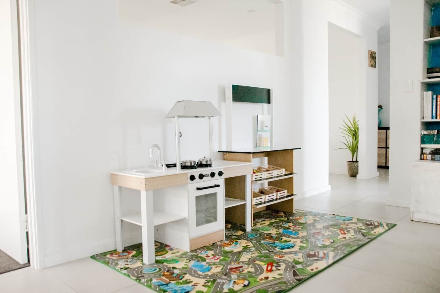 A child's play kitchen with a toy stove, sink, and shelves, a colorful play mat, and a bookshelf in the background.