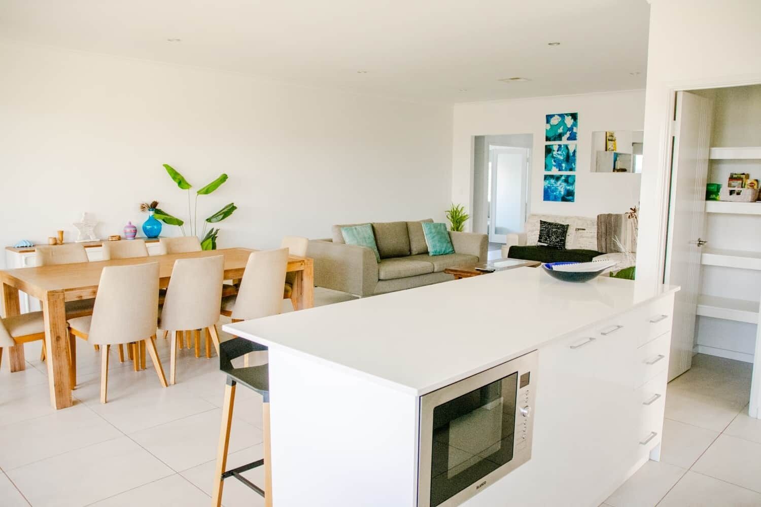 Cozinha moderna com uma ilha central, sala de jantar com mesa e cadeiras de madeira, e sala de estar com sofá e almofadas decorativas.