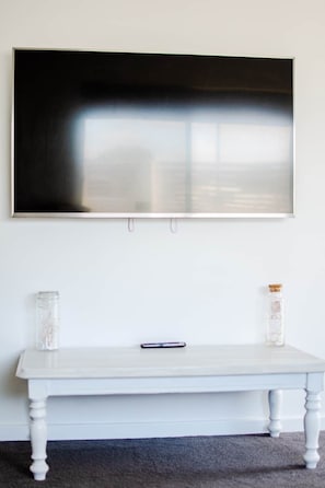 Una pared blanca con una obra de arte en un marco negro grande, una mesa blanca con dos frascos de vidrio y un control remoto sobre la mesa.