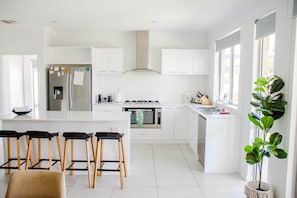A modern kitchen with white cabinetry, a central island with bar stools, a built-in oven, and a large window. - Swell House (Dunsborough)