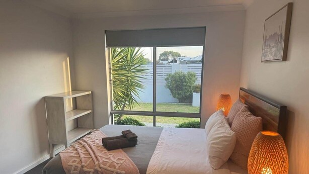 Une chambre à coucher avec un lit, une lampe de chevet et une vue sur la verdure et un bâtiment, le tout visible par une fenêtre.