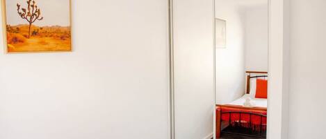 A room with a sliding door leading to a bed with red bedding, a small table, and a chair.