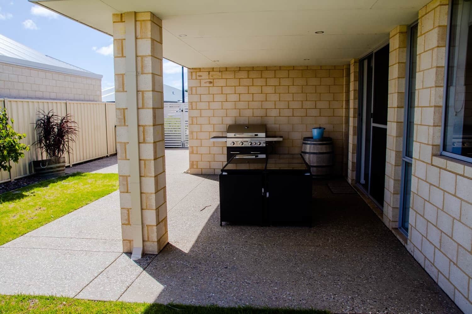 Un patio couvert comprenant un barbecue, un tonneau pour l’eau et un sol en béton.