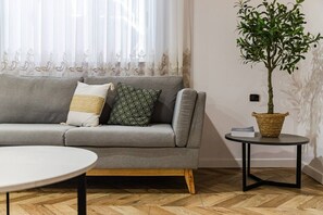 Een grijze bank met patroon kussens, een ronde witte tafel, een klein rond bijzettafeltje met een plant, en een visgraat houten vloer.