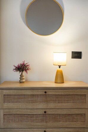 Een nachtkastje met een lamp, een vaas met bloemen en een spiegel aan de muur.
