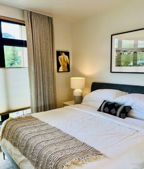 A hotel room with a bed, a nightstand, a lamp, a framed picture, and a window with curtains.