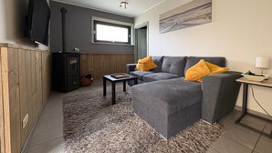 Una sala de estar con un sofá gris, paneles de madera, un televisor, una ventana y un cuadro en la pared.