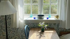 Una habitación con ventana, una mesa con un florero, una lámpara y papel tapiz estampado.