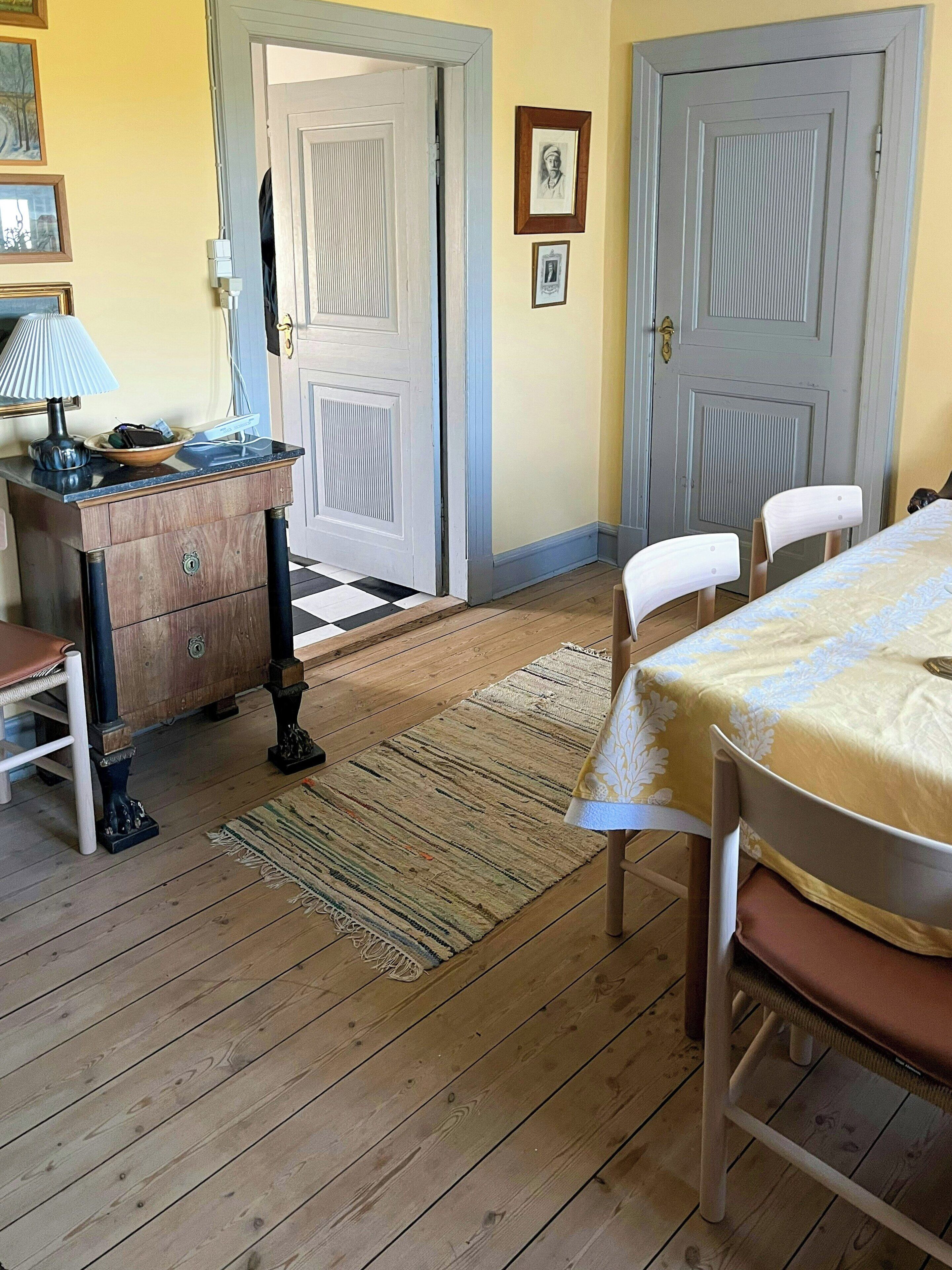 A room with a wooden floor, a dining table with chairs, a small wooden cabinet, and two doors.