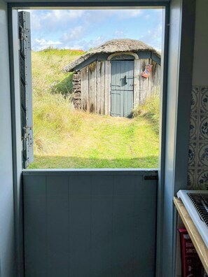 Blick durch eine Tür auf ein rustikales Holzgebäude mit einem Strohdach, einer blauen Tür und einem kleinen roten Briefkasten.
