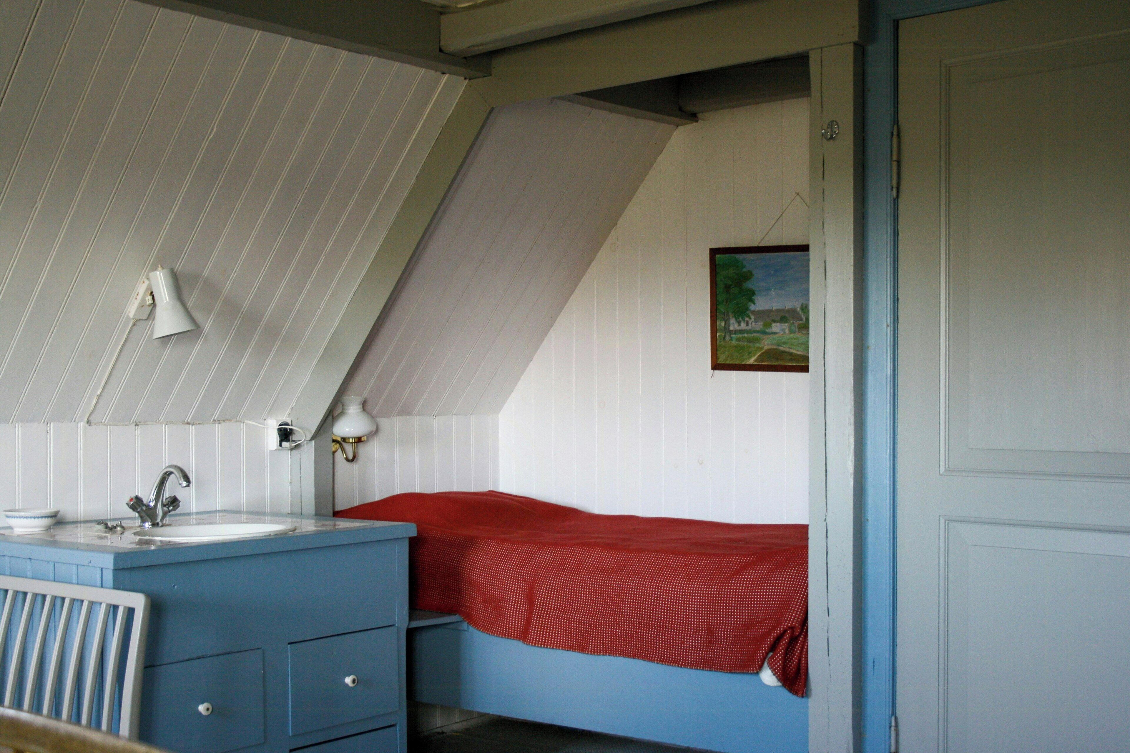 Ein Zimmer mit schrägen Wänden, einem Bett mit roter Decke, einer weißen Wand mit einem gerahmten Bild und einer blauen Kommode mit Schubladen.