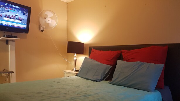 A bed with a blue bedspread, a nightstand with a lamp, a wall-mounted TV, and a standing fan. - Room-boutique Self Catering Guesthouse 4 (Johannesburg)