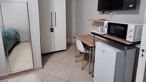 Een kleine kamer met een bed, een kledingkast, een bureau met stoel, een magnetron, een koelkast en een televisie aan de muur.