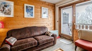 A cozy room with a brown sofa, wooden walls, and a large window with a view of a snowy landscape. - Le Nid de Châtel - Joli studio pour 4 avec balcon (Châtel)