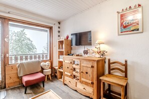 Ein gemütliches Zimmer mit einer hölzernen Sitzbank, einem Fernseher, einer Holzkommode und einem Fenster mit Blick auf Bäume.