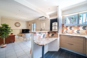 A modern kitchen with a built-in island, a dining area with a sofa and TV, and a large window with a view of palm trees. - L'Horizon Doré - 500m de la Plage (Saint-Cyprien)