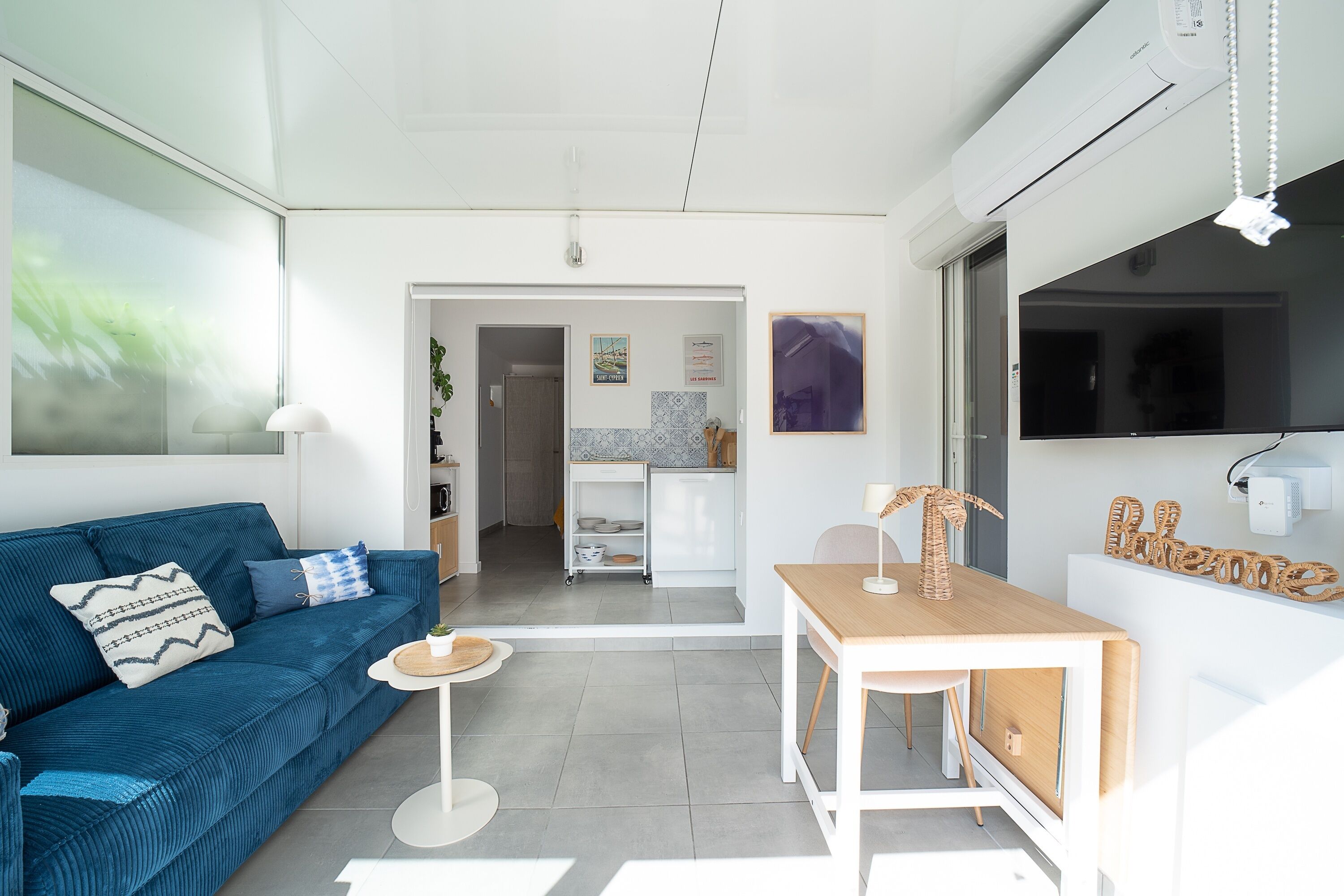 Ein modernes Wohnzimmer mit einem blauen Sofa, einem kleinen runden Tisch und einem Holzessttisch. Im Hintergrund ist eine Küchenzeile erkennbar.