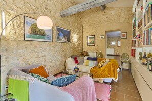 A cozy living room with a stone wall, a sofa with colorful cushions, a coffee table, and a bookshelf filled with books. - Villa Douceur - Avec piscine privée et sauna (Condom)