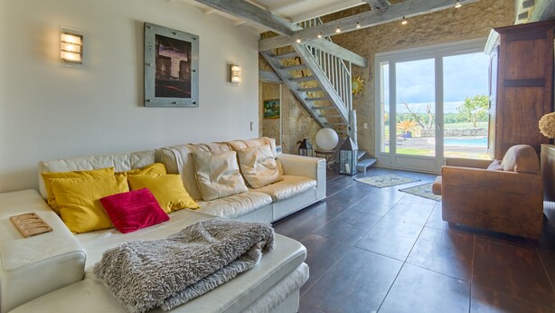 Amplia sala de estar con un sofá blanco, cojines amarillos y rojos, una manta gris y una silla de madera. Una escalera con barandilla de madera lleva a un balcón con vistas al exterior.