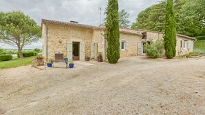 Una casa de piedra con camino de grava, dos cipreses altos y un banco con plantas en macetas.