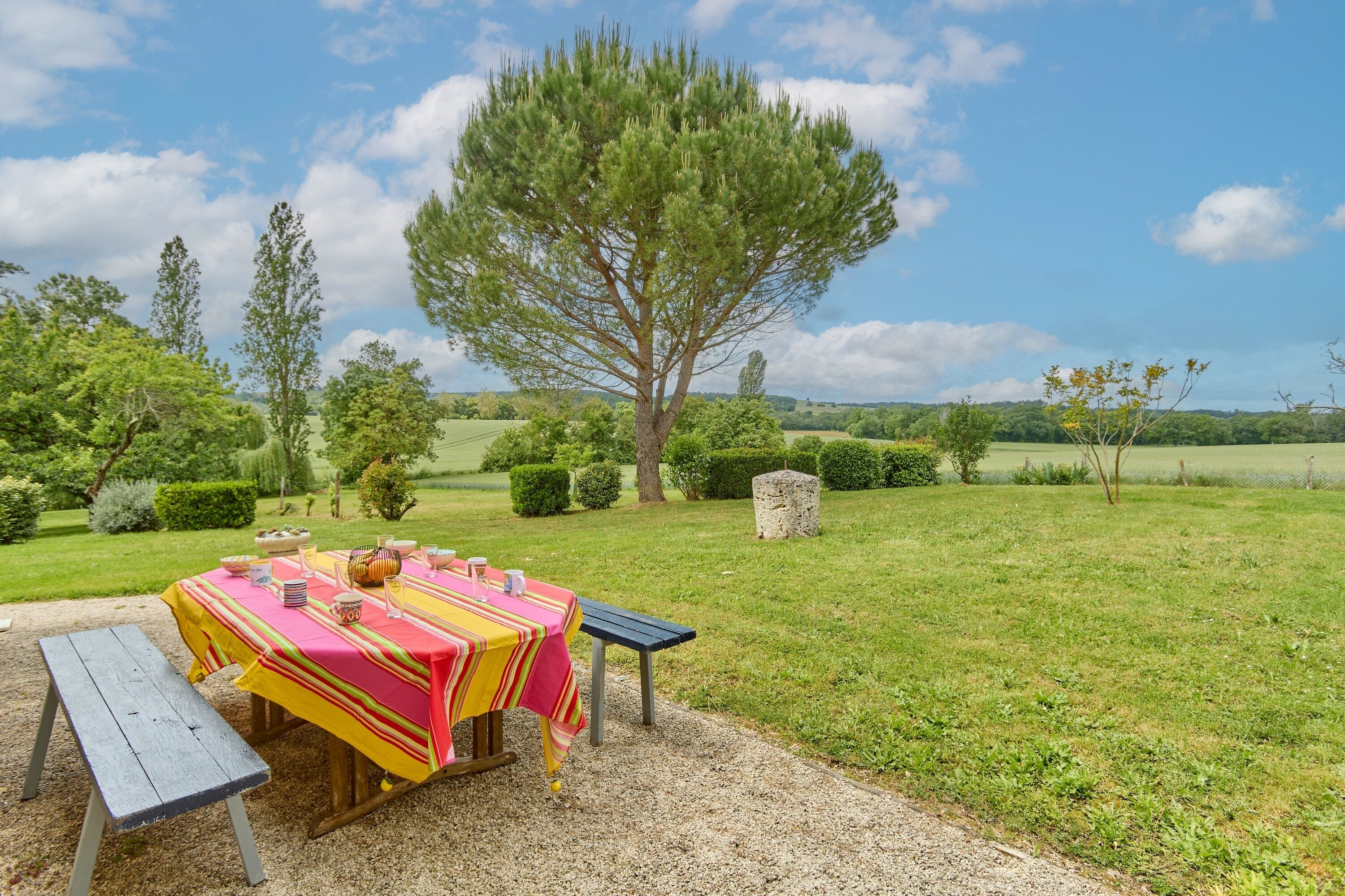 A picnic table with a striped tablecloth, a bench, and a stone structure in a grassy outdoor area with trees and a clear sky.
