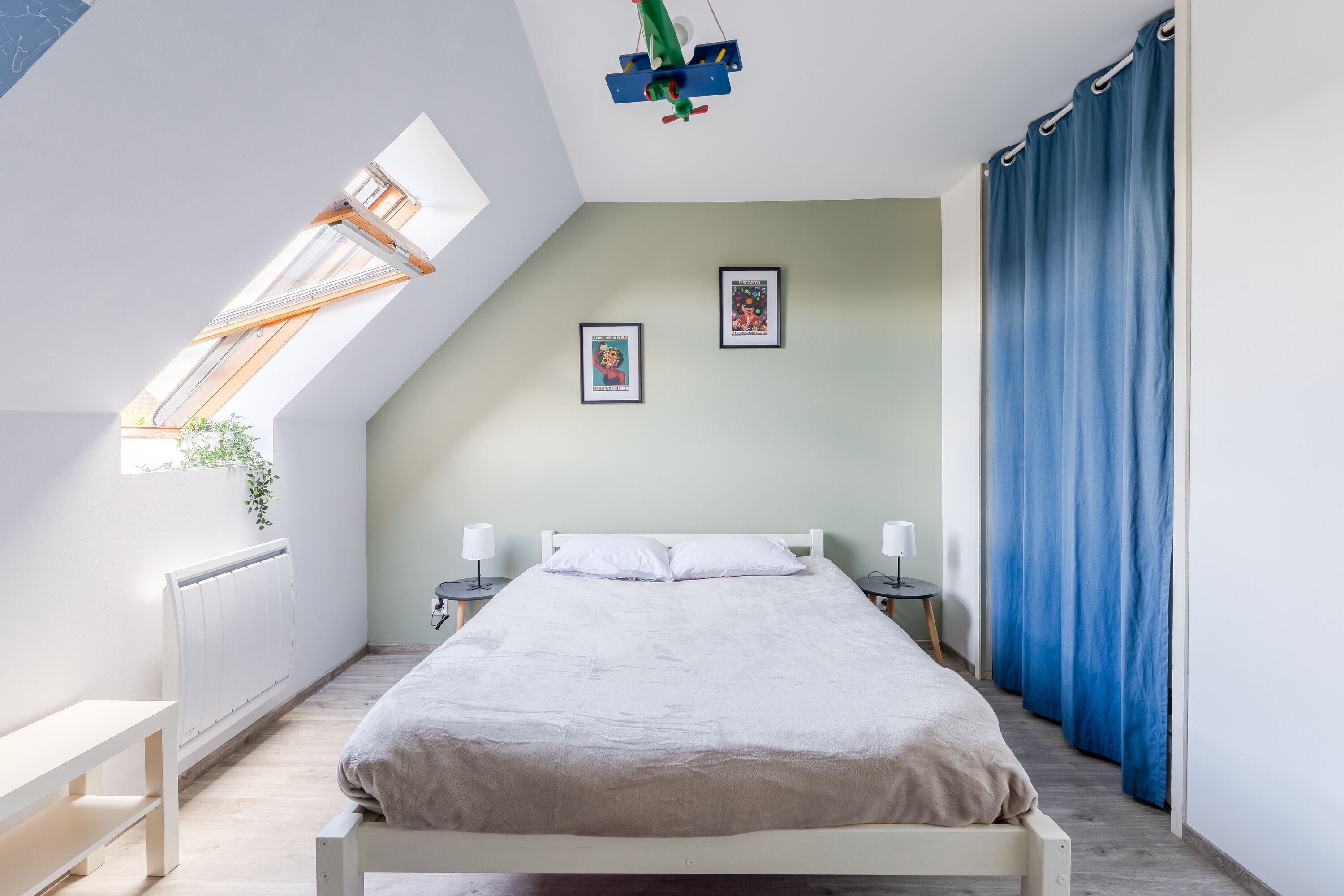Ein Schlafzimmer mit schrägverdeckter Decke, einem Bett, Nachttischen, einem Fenster mit Ausblick und einem blauen Vorhang.