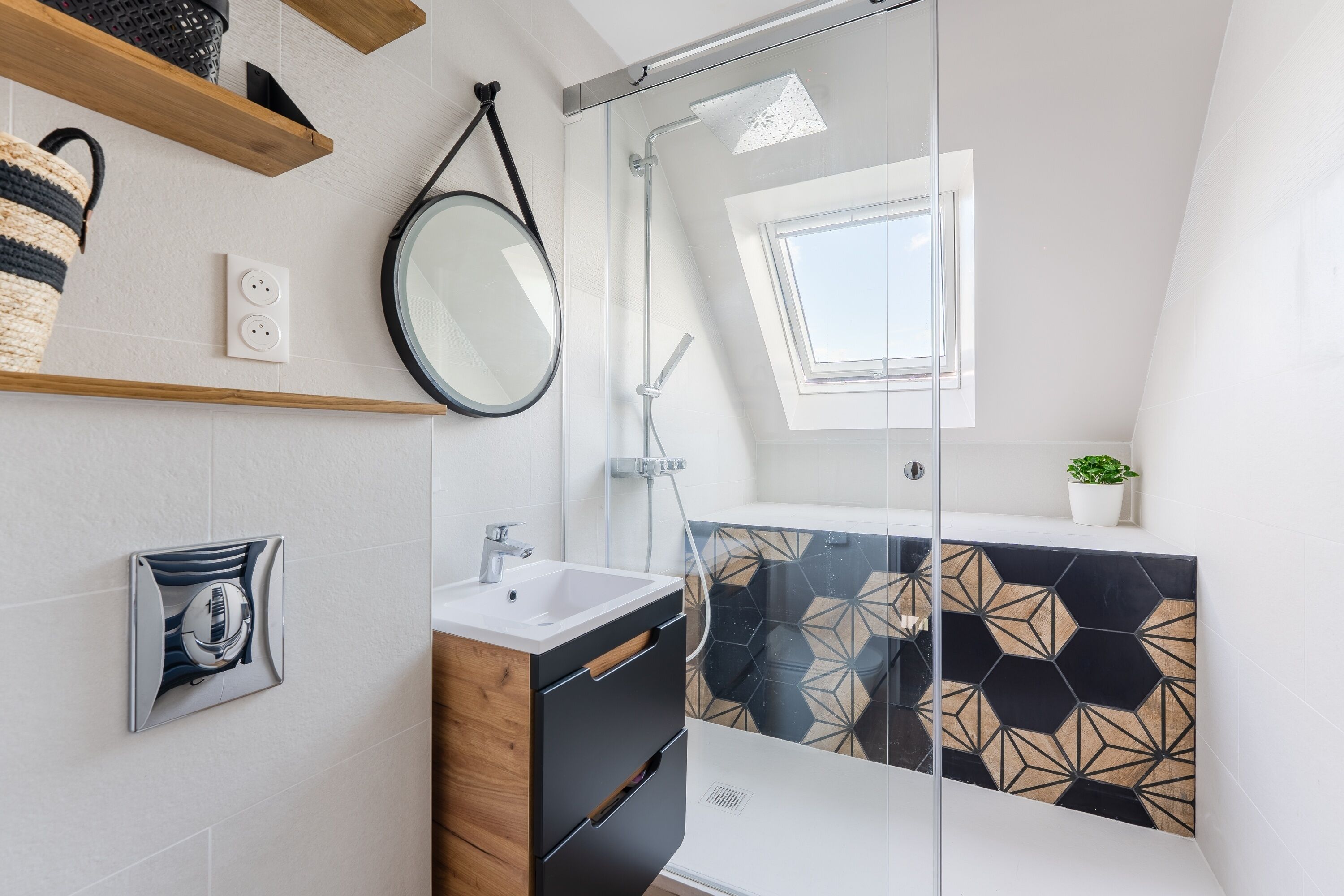 Une salle de bain moderne avec une douche à parois de verre, un miroir rond, un meuble-lavabo en bois et un revêtement mural géométrique en carreaux.