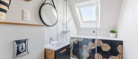 Un baño moderno con cabina de ducha de vidrio, espejo redondo, tocador de madera y salpicadero de azulejos geométricos.