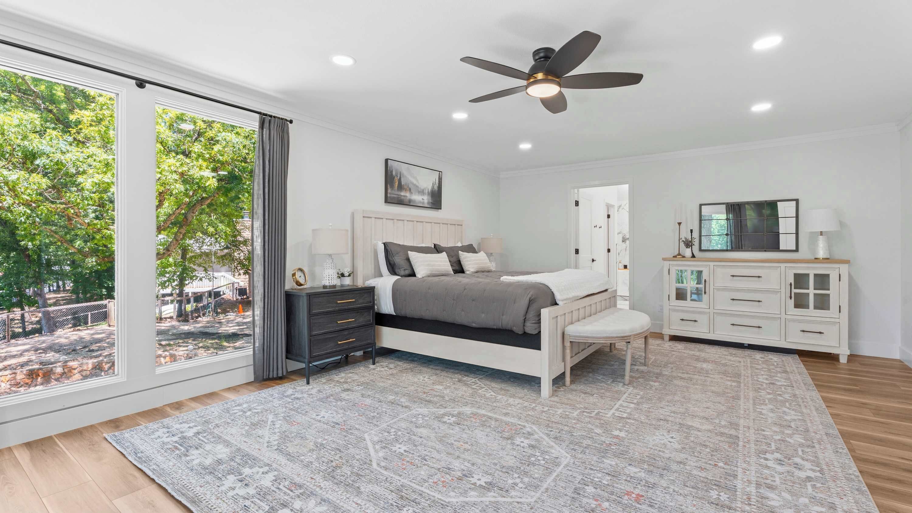 Une chambre à coucher comprenant un lit, des tables de chevet, un ventilateur de plafond, une grande fenêtre et une vue sur l’extérieur.