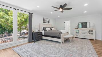 Une chambre à coucher comprenant un lit, des tables de chevet, un ventilateur de plafond, une grande fenêtre et une vue sur l’extérieur.