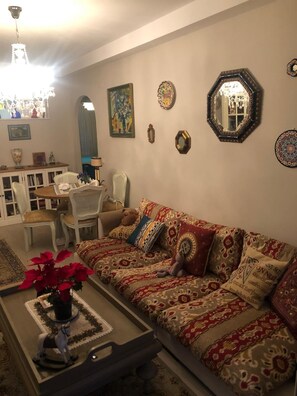 Una sala de estar acogedora con un sofá estampado, una mesita de centro con una bandeja decorativa y diversas decoraciones de pared, incluyendo un espejo y obras de arte enmarcadas.