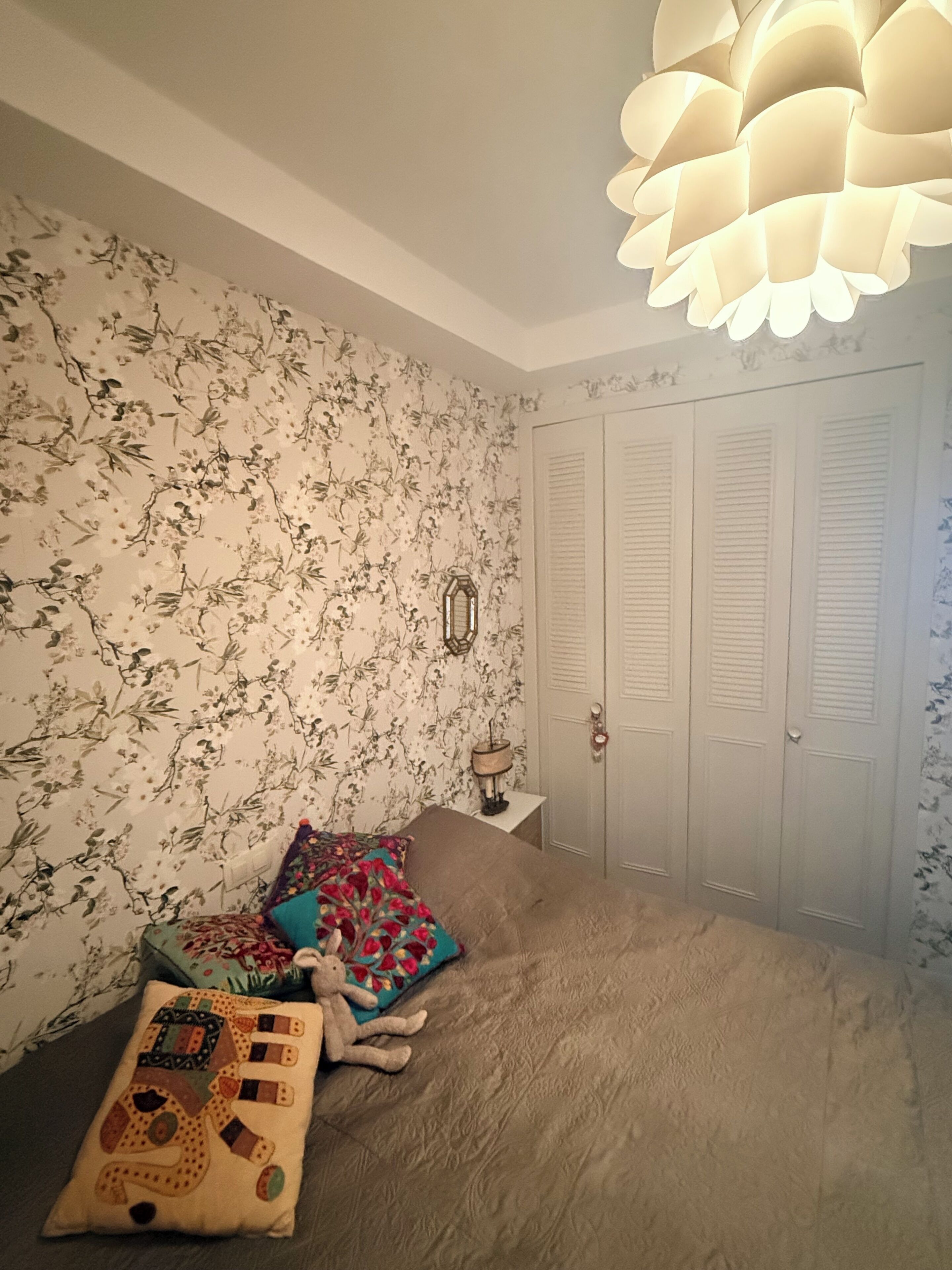 Une chambre à coucher avec un papier peint à motifs floraux, un lit avec de nombreux coussins, une table de chevet avec une lampe et une armoire à portes à persiennes.