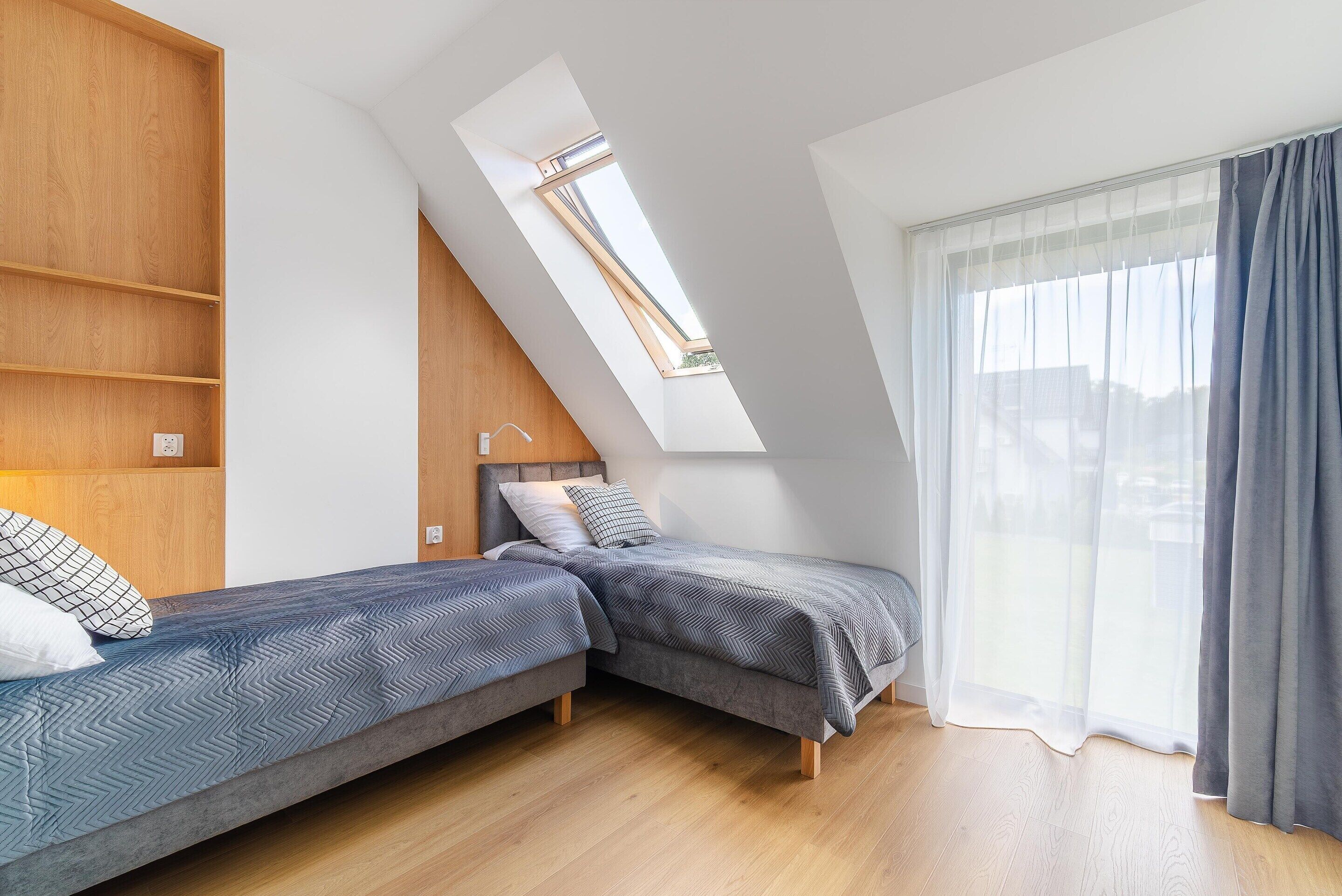A room with two beds, a skylight, a large window with curtains, and wooden flooring.