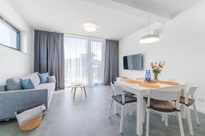 Ein modernes Wohnzimmer mit einer Couch, einem Esstisch, Stühlen, einem Fernseher und einem großen Fenster mit Vorhängen.