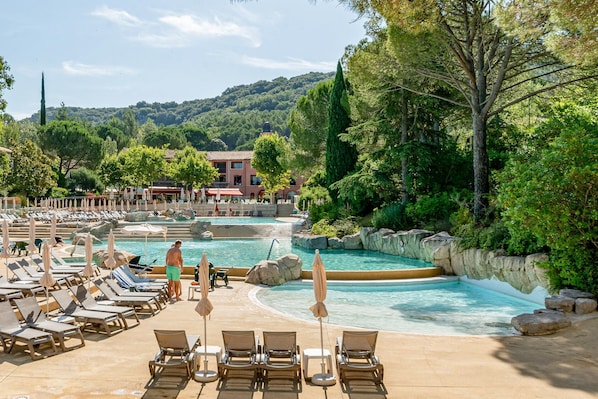 Un espace piscine avec des chaises longues et des parasols, entouré de verdure et avec un bùtiment en arriÚre-plan.