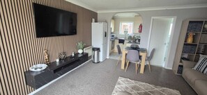 Una sala moderna con un televisor de pantalla plana, un comedor con sillas, una cocina al fondo y un sofá.