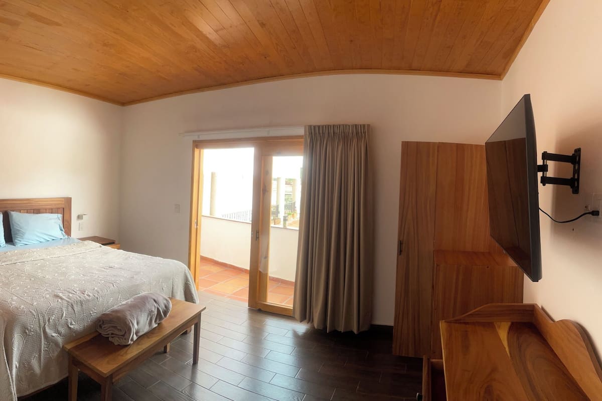 Un dormitorio con cama, mesita de noche, banqueta, techo de madera y televisor montado en la pared.