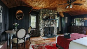 Una habitación con chimenea de piedra, una cama con manta roja, una mesa de comedor con sillas y un ventilador de techo.