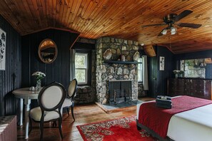 A room with a stone fireplace, a bed with a red blanket, a dining table with chairs, and a ceiling fan. - Swannanoa Guest House: Stunnning Mountain Views (Afton)