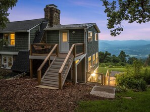 Ein zweistöckiges Haus mit einem Steinsschornstein, einer Holzterrasse und Blick auf Berge.
