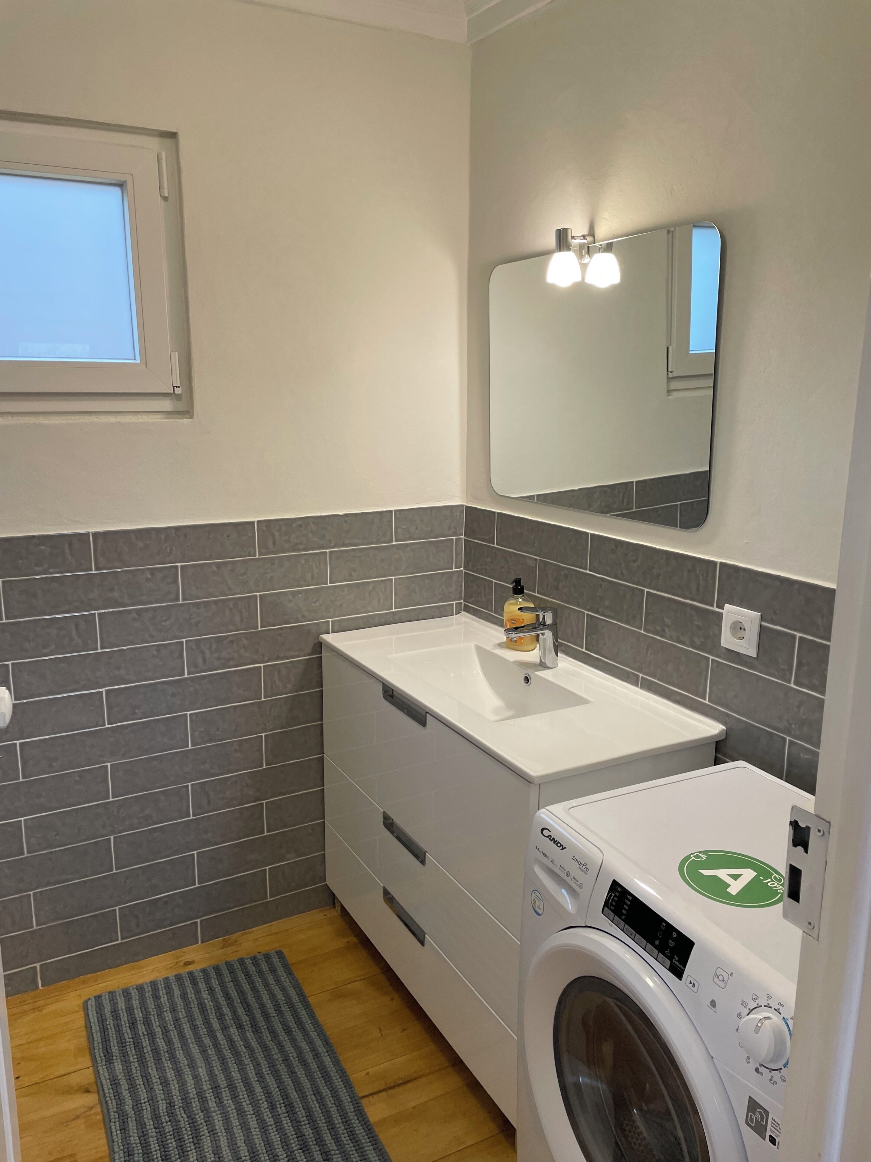 Un baño pequeño con lavamanos blanco, un espejo, lavadora y ventana.