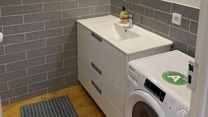 Ein kleines Badezimmer mit einem weißen Waschbecken, einem Spiegel, einer Waschmaschine und einem Fenster.