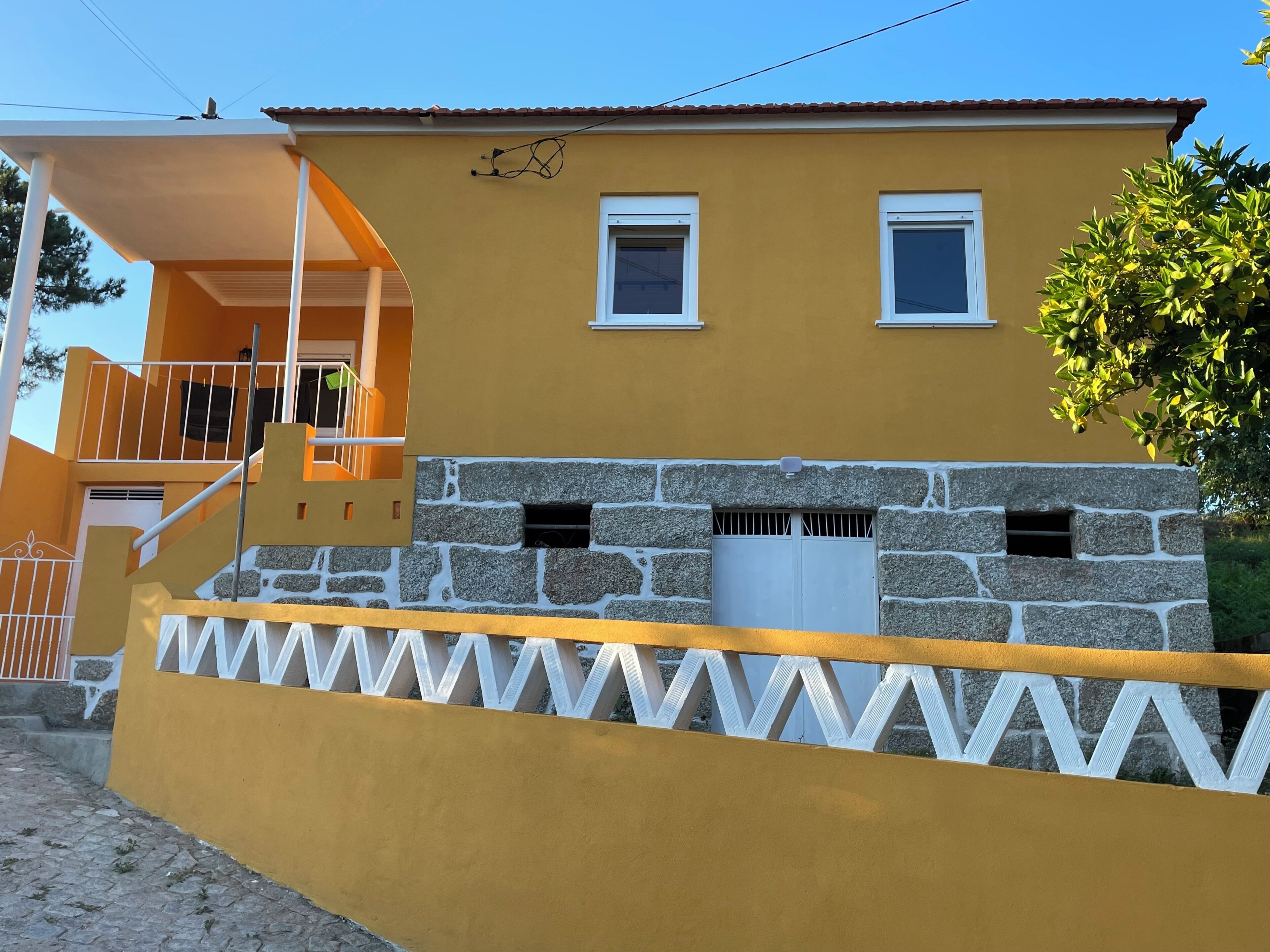 Un edificio amarillo con dos ventanas, base de piedra y valla blanca.