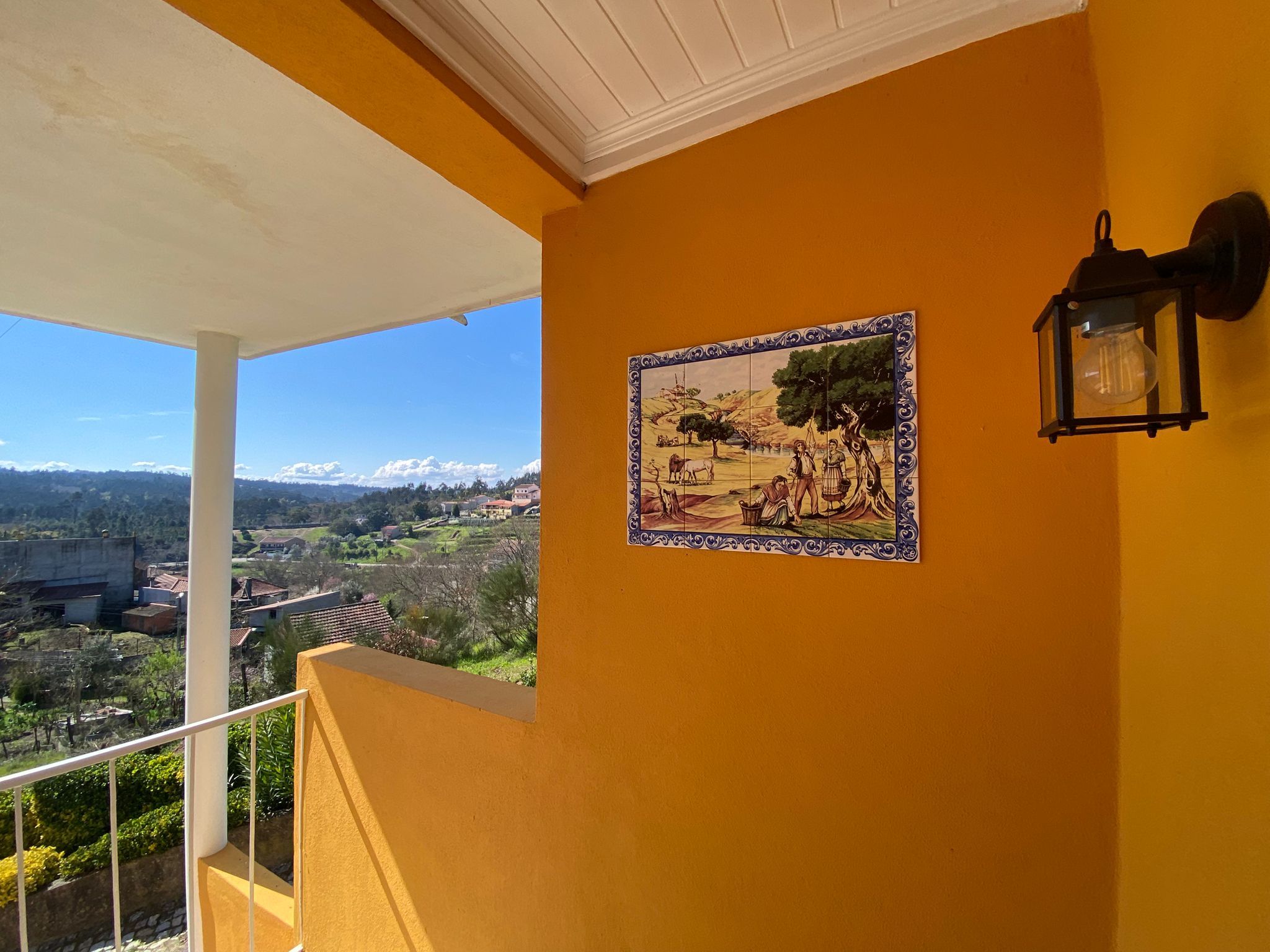 Un balcón con vistas a un paisaje rural, una obra de arte de azulejos y una lámpara fijada a la pared.