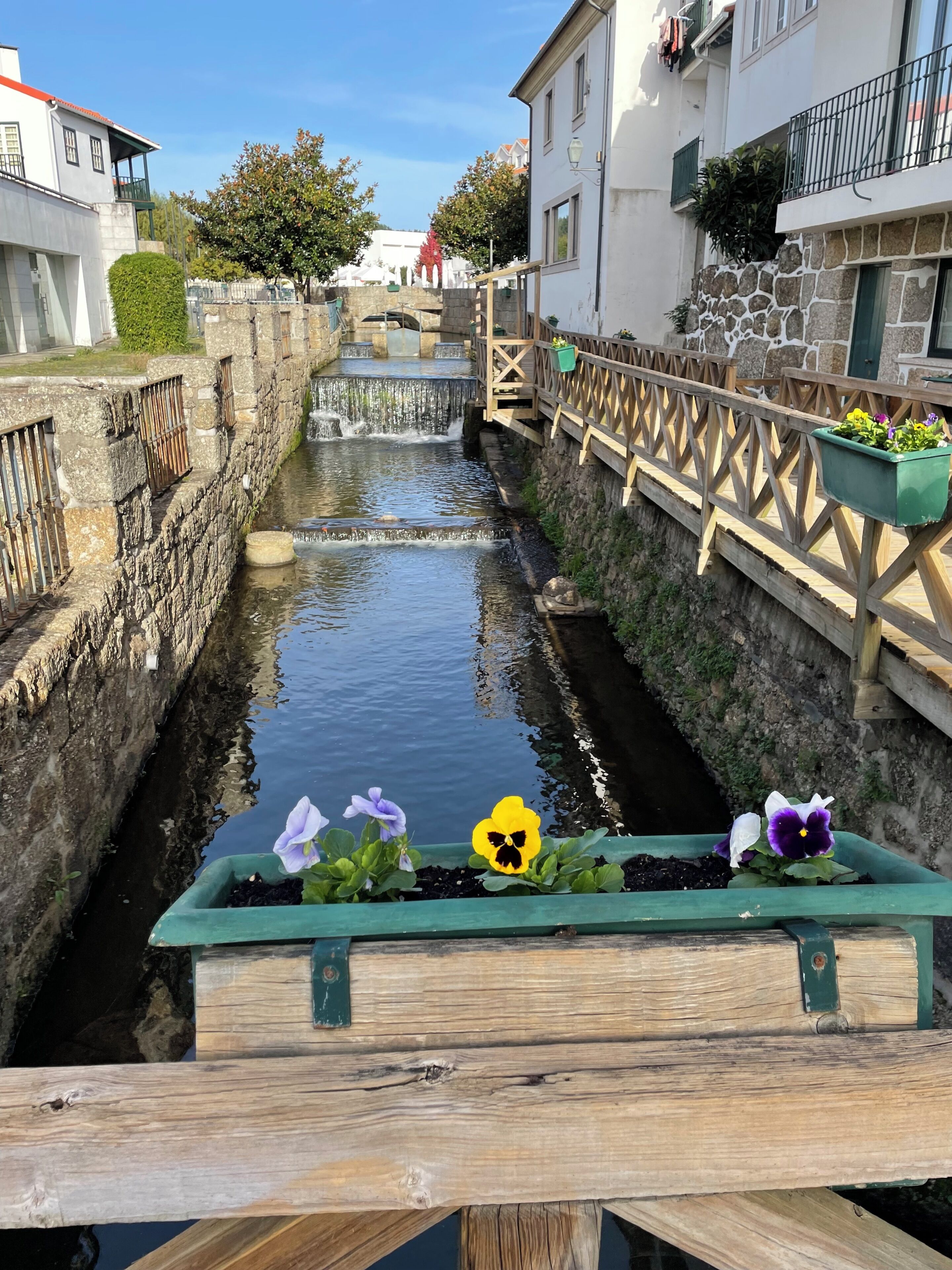 En smal vandkanal med stenvægge, en træbro og blomsterkasser.