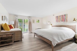 A bedroom with a bed, a desk, and a chair. There is a window with curtains, a wall-mounted lamp, and a colorful artwork.