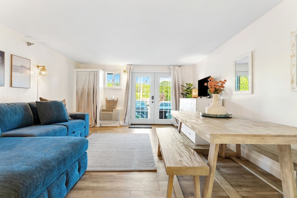 A living room with a blue sofa, a wooden dining table, and a view through a glass door.