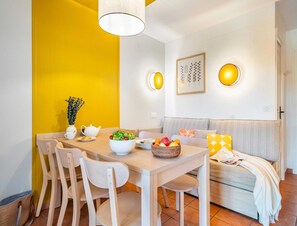 A dining area with a wooden table, white chairs, a yellow accent wall, and a sofa with patterned cushions. - Family Suite With Garden Terrace (Mandelieu-La-Napoule)