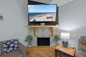 A living room with a TV displaying a beach scene, a fireplace with patterned tiles, and two potted plants. - 4BR Foley Home Near OWA and Beaches (Foley)